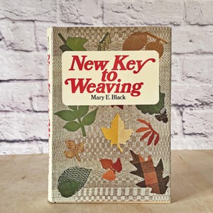 Peut inclure: Un livre relié intitulé New Key to Weaving par Mary E. Black. La couverture présente un fond en tissu tissé beige avec un motif de diverses feuilles d'automne colorées dans des tons de vert, jaune, orange, marron et rouge sur le devant.