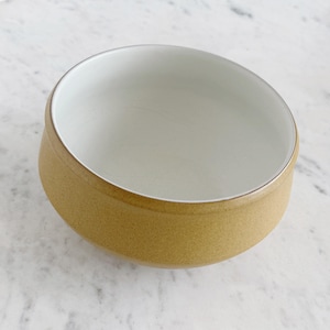 May include: A small, round bowl with a light brown exterior and a white interior. The bowl has a simple, minimalist design, with a smooth, matte finish. The rim of the bowl is slightly angled, giving it a modern look. The bowl is empty.