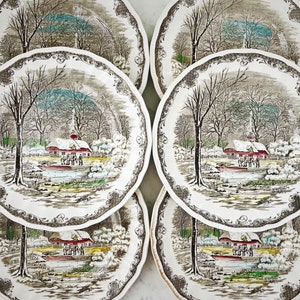 May include: Six vintage ceramic dinner plates with a winter scene featuring a red farmhouse, snow-covered trees, and a pond. The plates have a brown and white floral border.