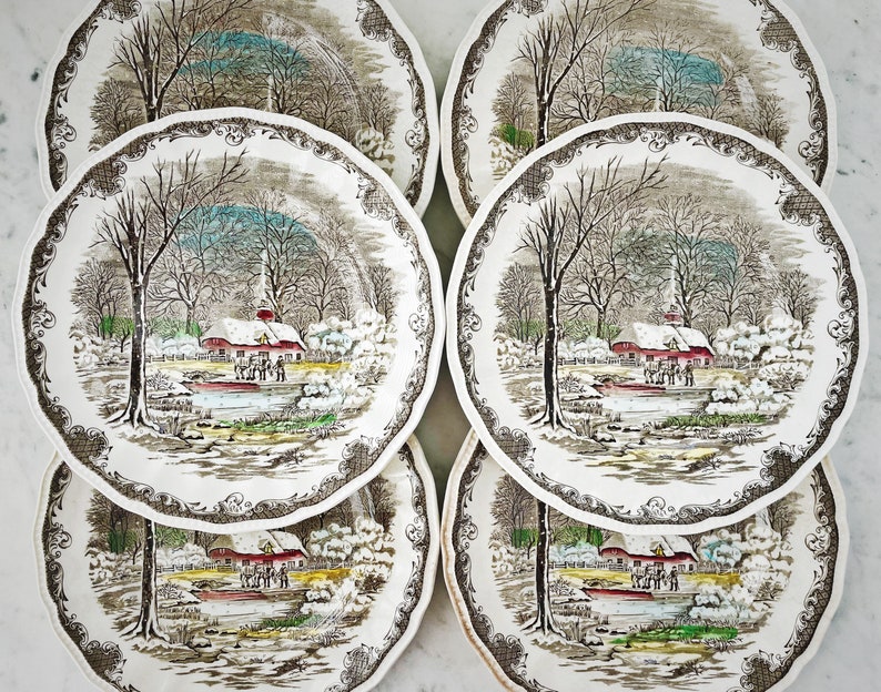 May include: Six vintage ceramic dinner plates with a winter scene featuring a red farmhouse, snow-covered trees, and a pond. The plates have a brown and white floral border.