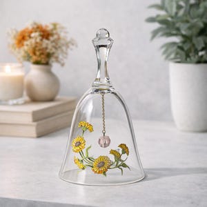 May include: A clear glass bell-shaped cloche with a decorative floral design. The bell features yellow and orange flowers with green stems. A small pink crystal hangs from a chain inside the bell. The bell has a clear handle.