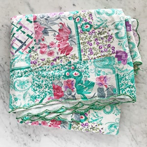 May include: A patchwork quilt with a white background and a variety of floral patterns in shades of pink, green, and purple. The quilt has a scalloped edge and is folded in half.