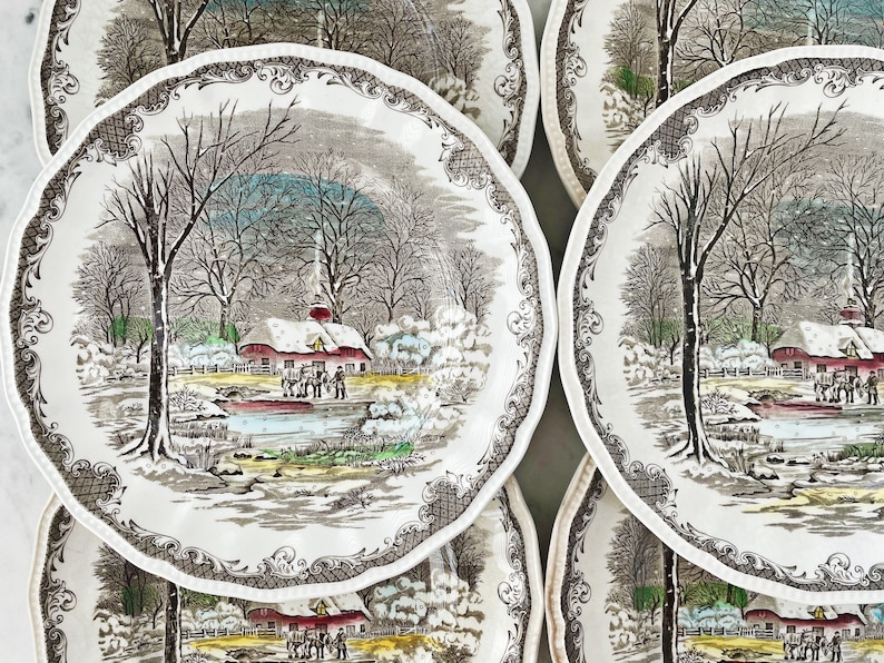 May include: Set of six vintage ceramic plates featuring a winter scene with a red farmhouse, snow-covered trees, and a frozen pond. The plates have a brown and white floral border.
