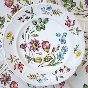 May include: White ceramic plates with a scalloped edge, decorated with a floral pattern. The design includes purple, red, yellow, and blue flowers with green leaves. The plates have a classic, elegant aesthetic.
