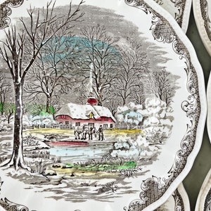 May include: A vintage ceramic plate with a winter scene featuring a red farmhouse, snow-covered trees, and figures ice skating on a frozen pond. The plate has a brown and white floral border.