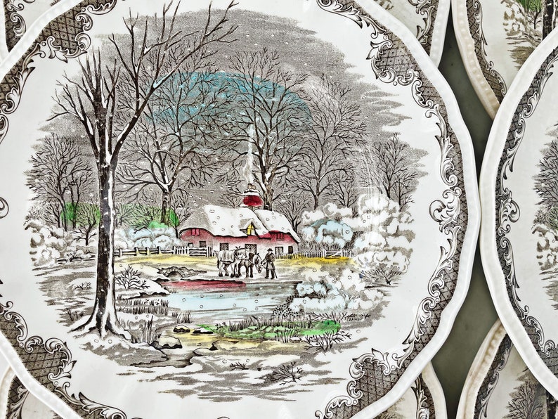 May include: A vintage ceramic plate with a winter scene featuring a red farmhouse, snow-covered trees, and figures ice skating on a frozen pond. The plate has a brown and white floral border.