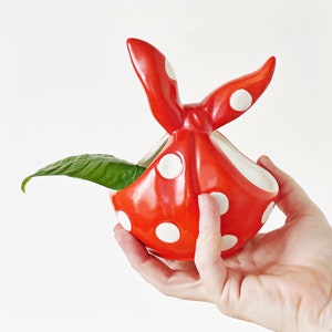 Op de afbeelding: Rode keramische plantenpot in de vorm van een bandana met witte stippen. De pot heeft een kleine opening waar een plant uit kan groeien.
