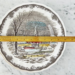 May include: A vintage ceramic dinner plate with a winter scene featuring a farmhouse, trees, and a frozen pond. The plate has a brown and white border with a repeating pattern.