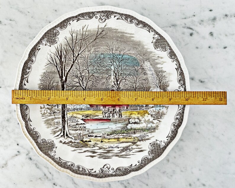 May include: A vintage ceramic dinner plate with a winter scene featuring a farmhouse, trees, and a frozen pond. The plate has a brown and white border with a repeating pattern.
