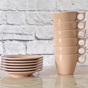 Peut inclure: Six tasses pêche empilées avec des poignées originales, à côté d'une pile de soucoupes assorties. Les tasses sont empilées verticalement, et les soucoupes sont empilées horizontalement. Le fond est un mur de briques blanches.