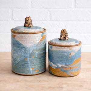 May include: Two ceramic jars with lids, glazed in blue and white with orange highlights. Each lid is adorned with a small cat figurine. The cylindrical jars have a handmade appearance, suggesting a rustic, artisanal style.