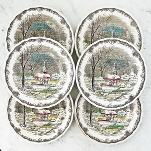 May include: Set of six vintage ceramic dinner plates featuring a winter scene with a red farmhouse, trees, and snow. The plates have a brown and white floral border.