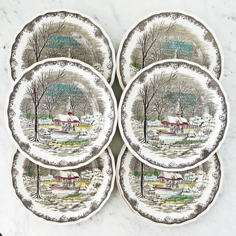 May include: Set of six vintage ceramic dinner plates featuring a winter scene with a red farmhouse, trees, and snow. The plates have a brown and white floral border.