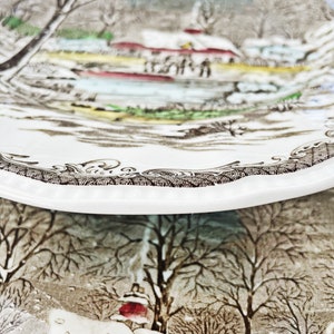 May include: A vintage ceramic plate with a winter scene featuring a small village, trees, and a river. The plate has a brown and white design with a scalloped edge.