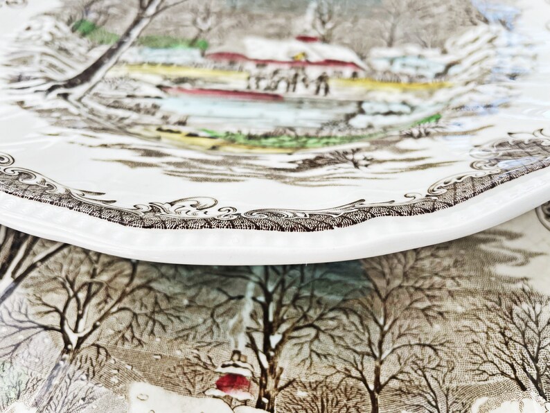 May include: A vintage ceramic plate with a winter scene featuring a small village, trees, and a river. The plate has a brown and white design with a scalloped edge.