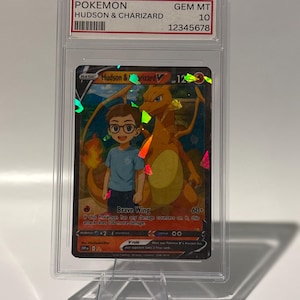 May include: A graded Pok&eacute;mon card featuring "Hudson & Charizard" with a GEM MT 10 grade. The card showcases a vibrant illustration of a boy and a Charizard, with the "Brave Wing" attack text visible. The card is encased in a clear protective holder.