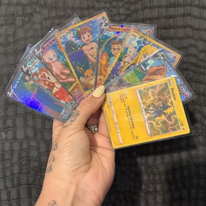 May include: A collection of holographic trading cards featuring various characters and the text "Ario & Me", "Sam & Umbrum", "Hudson & Charle", and "Minnie V". The cards are in protective sleeves, with a yellow card featuring a shadow graphic.