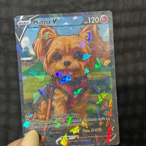 May include: A holographic trading card featuring a cartoon Yorkshire Terrier named Minnie V. The card has a blue and green background with the text "Starlight Hug" and "120 HP". The card is held by a person.