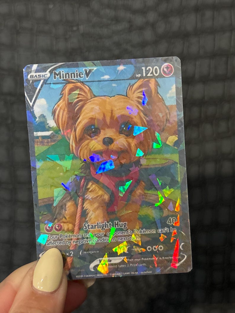 May include: A holographic trading card featuring a cartoon Yorkshire Terrier named Minnie V. The card has a blue and green background with the text "Starlight Hug" and "120 HP". The card is held by a person.