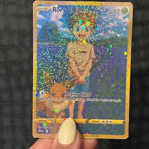 May include: A holographic trading card featuring "Rory & Eevee." The card depicts a boy and a cartoon animal in a field of flowers. The text "Helping Hand" is visible, along with other game-related symbols and numbers. The card has a gold border.