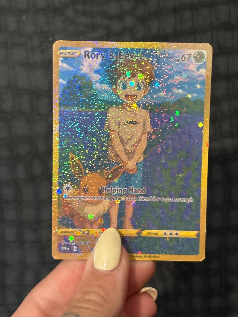 May include: A holographic trading card featuring "Rory & Eevee." The card depicts a boy and a cartoon animal in a field of flowers. The text "Helping Hand" is visible, along with other game-related symbols and numbers. The card has a gold border.