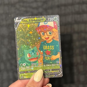 May include: A holographic Pok&eacute;mon trading card featuring "Elliott & Bulbasaur V." The card has a green and blue color scheme with the word "GRASS" on the character's shirt. The card has a holographic background with stars and sparkles.