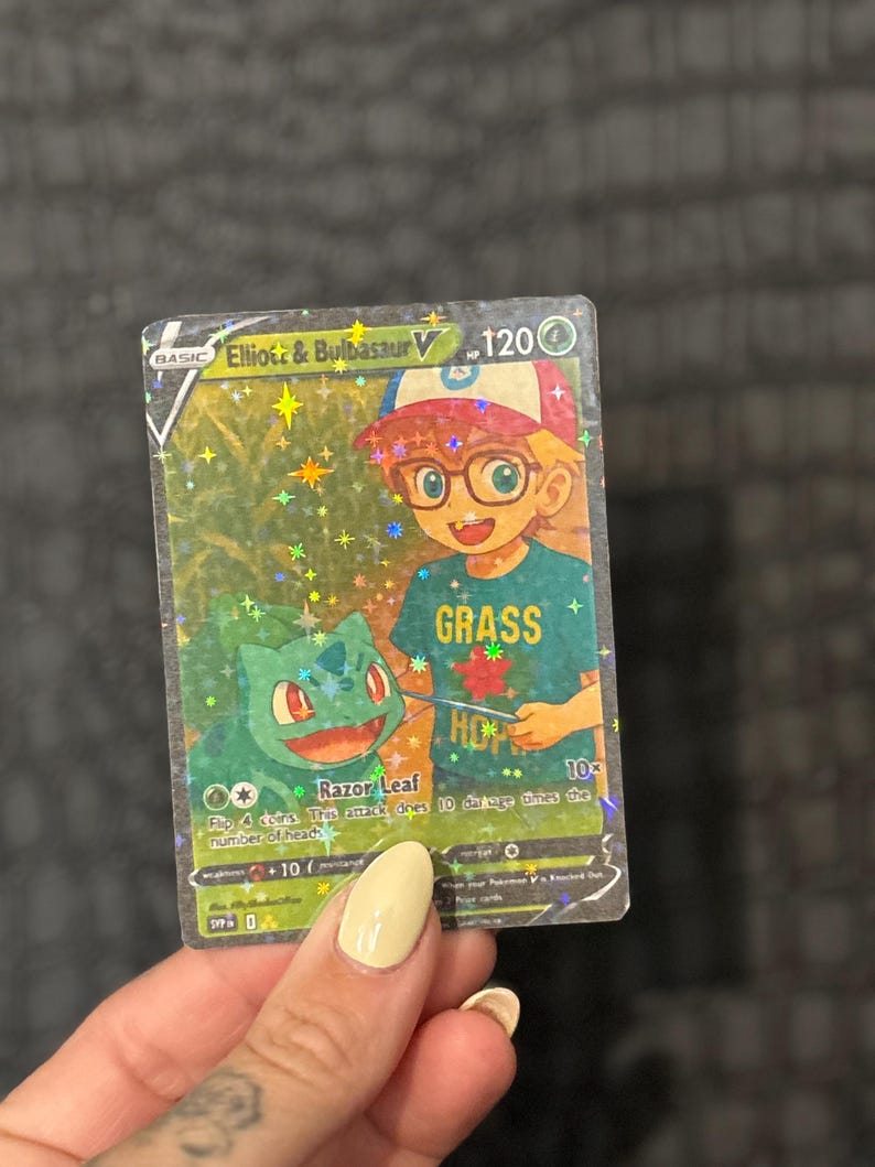 May include: A holographic Pok&eacute;mon trading card featuring "Elliott & Bulbasaur V." The card has a green and blue color scheme with the word "GRASS" on the character's shirt. The card has a holographic background with stars and sparkles.