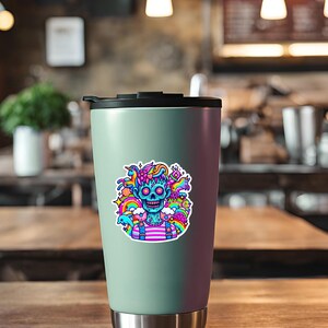 May include: A teal travel mug with a sticker of a blue skull with rainbow hair and a pink and white striped shirt. The skull is surrounded by colorful doodles, including a rainbow, stars, and a unicorn.