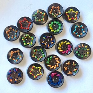 May include: A collection of 17 wooden buttons with colorful embroidery. The buttons are round and feature various designs, including stars, circles, and swirls. The buttons are all different colors, including red, blue, green, yellow, and orange.
