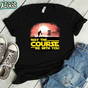 May include: Black t-shirt with a yellow graphic of a disc golf player throwing a disc. The graphic has a sunset background and the text "May the Course Be With You".