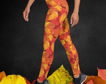 May include: A pair of leggings with a colorful autumn leaf print. The leggings are orange, yellow, and red with a pattern of leaves. The leggings are worn by a person standing on a bed of autumn leaves.