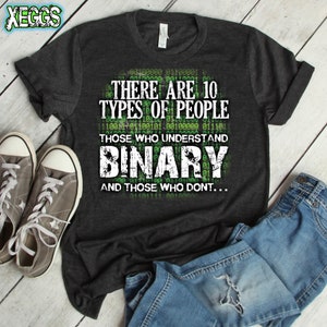 May include: A dark grey t-shirt with the text "THERE ARE 10 TYPES OF PEOPLE" and "THOSE WHO UNDERSTAND BINARY AND THOSE WHO DONT..." in white. The shirt is next to grey trainers and blue jeans.