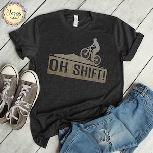 May include: A dark heather grey t-shirt with a distressed brown graphic of a mountain biker riding a bicycle with the text "OH SHIFT!" printed on it.
