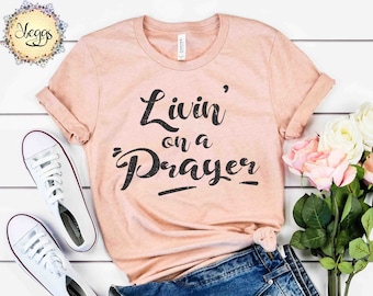 Peut inclure: Un t-shirt couleur pêche avec le texte "Livin' on a Prayer" en lettres noires. Le t-shirt est posé sur une surface blanche avec une paire de baskets blanches et un short en jean bleu.