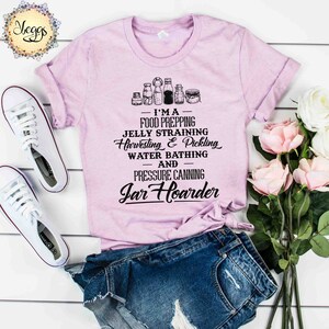 Puede incluir: Una camiseta rosa con un gr&aacute;fico negro que dice "Soy una preparadora de alimentos, una coladora de jalea, una cosechadora y una encurtidora, una ba&ntilde;adora de agua y una enlatadora a presi&oacute;n, una acaparadora de tarros". El gr&aacute;fico tambi&eacute;n incluye una fila de tarros.
