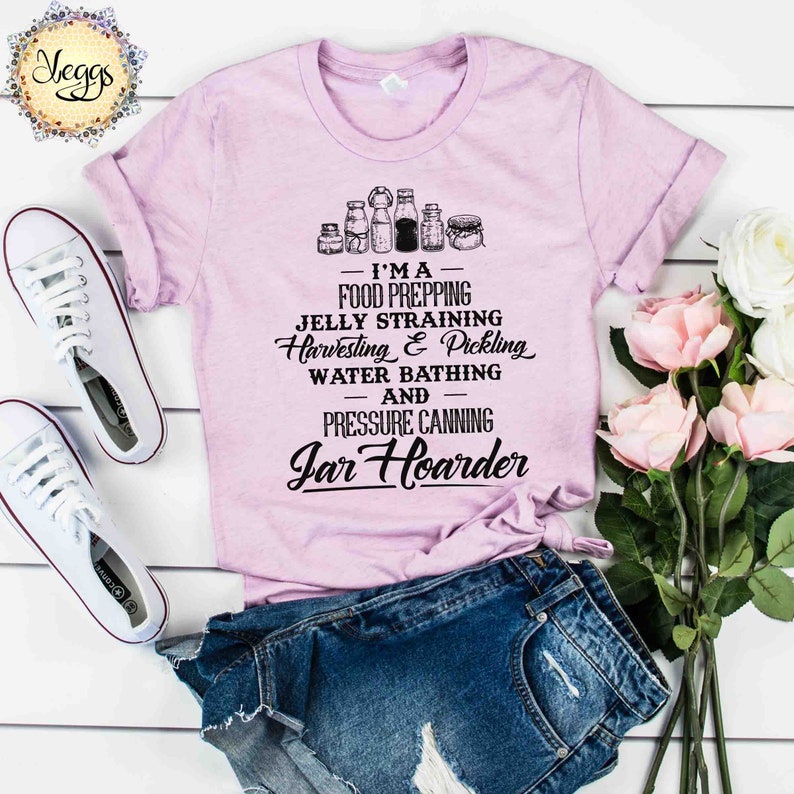 Puede incluir: Una camiseta rosa con un gr&aacute;fico negro que dice "Soy una preparadora de alimentos, una coladora de jalea, una cosechadora y una encurtidora, una ba&ntilde;adora de agua y una enlatadora a presi&oacute;n, una acaparadora de tarros". El gr&aacute;fico tambi&eacute;n incluye una fila de tarros.