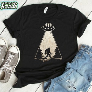 May include: Black t-shirt featuring a Bigfoot graphic being beamed up by a UFO. The design includes a forest silhouette and two aliens in the UFO. The shirt is paired with gray sneakers and blue jeans.