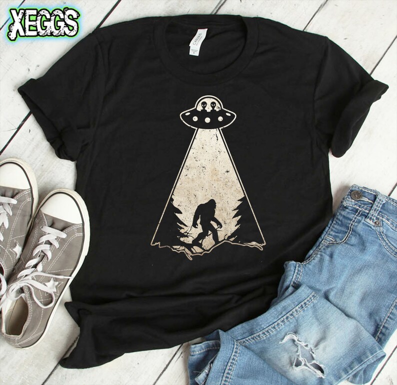 May include: Black t-shirt featuring a Bigfoot graphic being beamed up by a UFO. The design includes a forest silhouette and two aliens in the UFO. The shirt is paired with gray sneakers and blue jeans.