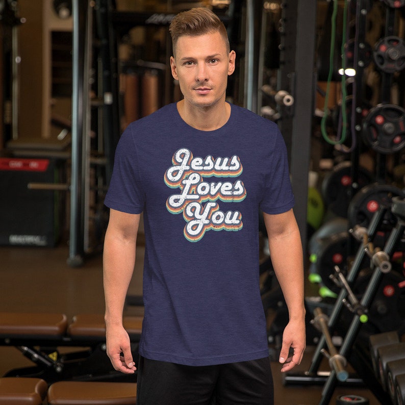 Puede incluir: Una camiseta azul marino con un gr&aacute;fico de estilo retro que dice "Jesus Loves You" en letras coloridas y descoloridas.