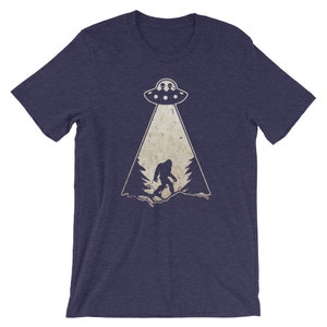 May include: Navy blue t-shirt featuring a Bigfoot graphic being abducted by a UFO. The graphic is in a vintage style with a beige color scheme. The t-shirt is made of a soft, comfortable material.