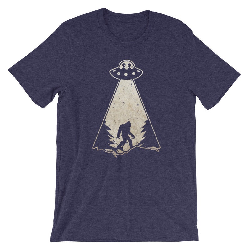 May include: Navy blue t-shirt featuring a Bigfoot graphic being abducted by a UFO. The graphic is in a vintage style with a beige color scheme. The t-shirt is made of a soft, comfortable material.