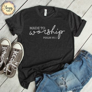 May include: A dark gray t-shirt with the text "MADE TO worship PSALM 95:1" in white lettering. The shirt is laid flat on a wooden surface with a pair of gray sneakers and blue jeans.
