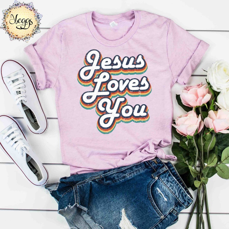 Puede incluir: Una camiseta rosa con el texto "Jesus Loves You" en una fuente retro arco&iacute;ris.