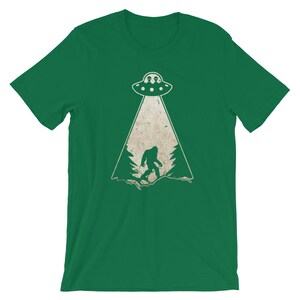 May include: Green t-shirt featuring a distressed graphic of a UFO beaming down on a Bigfoot figure in a forest. The UFO has two alien figures inside. The design is in white and tan.