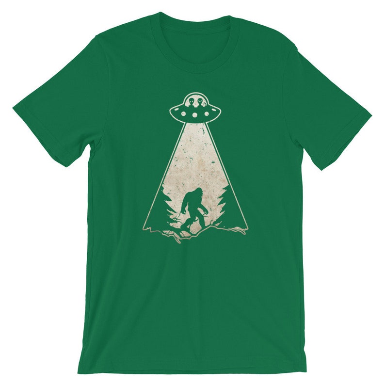 May include: Green t-shirt featuring a distressed graphic of a UFO beaming down on a Bigfoot figure in a forest. The UFO has two alien figures inside. The design is in white and tan.