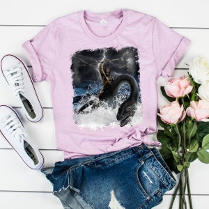 Puede incluir: Una camiseta rosa con un estampado gr&aacute;fico de un hombre de las cavernas montando un monstruo marino en un mar tormentoso. El gr&aacute;fico est&aacute; desgastado y tiene un borde blanco.