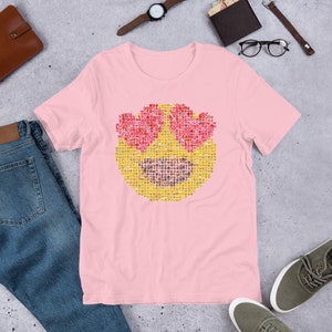 Puede incluir: Camiseta rosa claro con un gran emoji hecho de emojis m&aacute;s peque&ntilde;os. El emoji tiene ojos en forma de coraz&oacute;n y una boca sonriente. La camiseta est&aacute; hecha de un material suave.