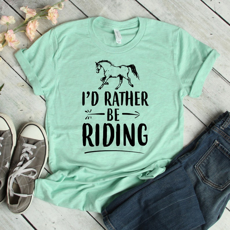 Puede incluir: Camiseta verde claro con el texto "I'd Rather Be Riding" y un dibujo de un caballo negro. La camiseta se combina con vaqueros azules y zapatillas grises, lo que sugiere un estilo informal de tem&aacute;tica ecuestre.
