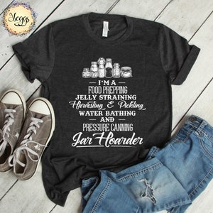 Puede incluir: Una camiseta gris oscuro con texto blanco que dice "I'MA FOOD PREPPING JELLY STRAINING Harvesting & Pickling WATER BATHING AND PRESSURE CANNING Jar Hoarder". La camiseta se muestra sobre una superficie de madera con un par de vaqueros y zapatillas.