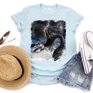 Puede incluir: Una camiseta azul claro con un estampado gr&aacute;fico de un hombre de las cavernas montando un monstruo marino en un mar tormentoso. El gr&aacute;fico est&aacute; desgastado y tiene un aspecto vintage.